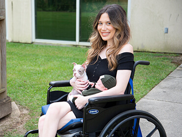 Young woman with long hair sitting in a wheelchair outside and holding a puppy and a baby.